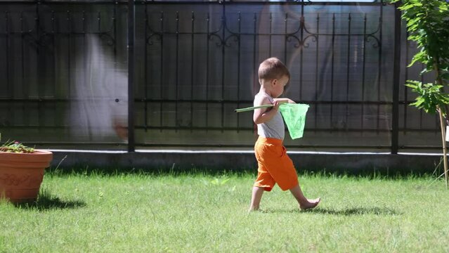 baby boy child walking playing with butterfly net in backyard outdoors sunny day. barefoot kid in orange shorts walking on green grass in garden with insect net trying to catch bugs