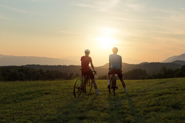 Professional cyclist couple taking a break on a green meadow and watching amazing mountain sunset, aerial shot.