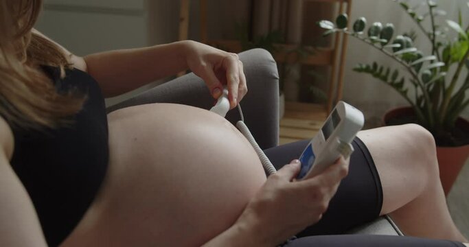 Relaxing pregnant woman use pocket ultrasound doppler stethoscope to check baby heart rate. The expectant loving mother caresses her baby in the tummy. Maternity prenatal care and pregnancy concept.
