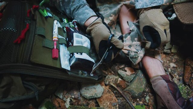 Close up hands military medic cuts the clothes of a wounded soldier with scissors to examine the body. Soldiers lying on ground. Blood around