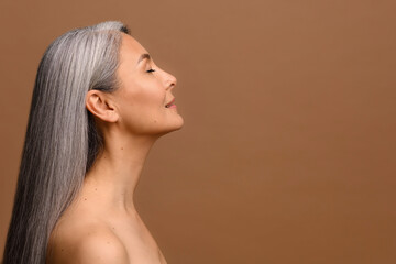 Side view at sensual naked middle-aged beautiful Asian woman with long grey hair, her eyes closed, body and face care concept. Charming mature lady stands in profile isolated on brown background