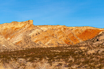 Fototapeta premium Overton, Nevada, USA - February 24, 2010: Valley of Fire. Orange-yellow mountain range forms a blet between blue sky and dry desert floor with brown shrubs.
