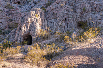 Overton, Nevada, USA - February 24, 2010: Valley of Fire. Dark no-light entrance to cave into gray mountain range behind dry desert floor with green shrubs. Partly lit by sun. - Powered by Adobe