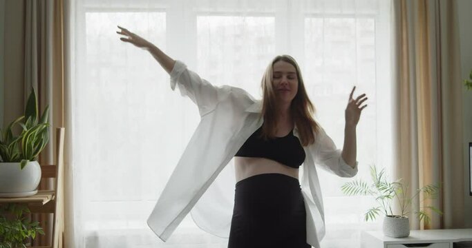 Happy cheerful pregnant young woman funny dancing near the window at home. Expectant loving mother relax at home listening to the music. Maternity prenatal care and woman pregnancy concept.