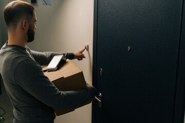 Back view of delivery man ringing doorbell of customer apartment holding cardboard box and...