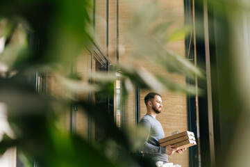 Remote view of delivery man holding boxes pizza and contactless payment POS terminal, standing in entrance hall of apartment or office building, by door by waiting to meet customer, looking away.