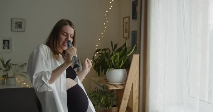Happy cheerful pregnant woman funny dancing and singing near the window at home. Expectant loving mother relax at home listening to the music. Maternity prenatal care and woman pregnancy concept.
