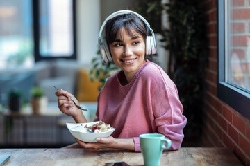 Beautiful young woman listening music with headphones while having healthy breakfast in the living...