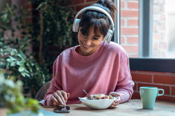 Beautiful young woman listening music with headphones while having healthy breakfast in the living...