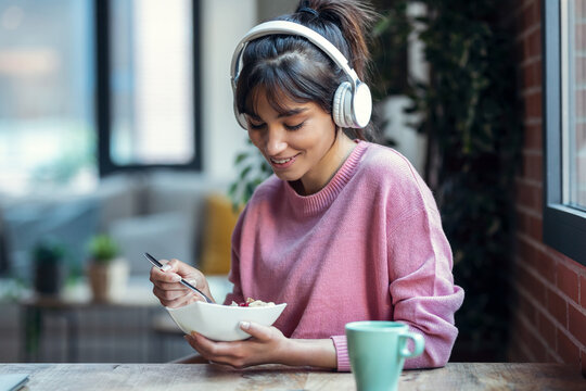 Beautiful young woman listening music with headphones while having healthy breakfast in the living room at home.
