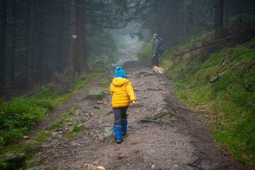 Mum and her little son go on a mountain trail in wet autumn weather. They are accompanied by a dog.