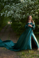 A beautiful model in a green silk dress with a long train on a path in a blooming apple orchard.