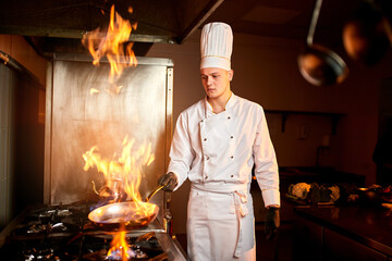 Сhef in the professional kitchen with a frying pan and a fire..Chef's hands hold iron Pan and preparing food on cooker.
