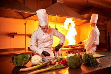 А team of professional chefs cook meals with frying pan and fire in the kitchen of restaurant.  Chief chef preparing dish using different food ingredients, vegetables, meat and fish
