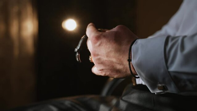 A Caucasian man holds an expensive Swiss watch with a leather strap and examines it. Successful man sitting in a room with a flashlight and thinking about something with a watch in his hand