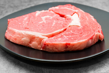 High angle studio shot of a raw beef rib eye steak on a black design plate standing on a grey concrete surface, with free copy space. Keto diet, warrior diet concept. Raw red meat ready for barbecue.