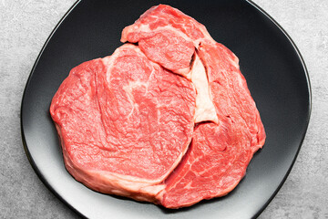 Top studio shot of a raw beef rib eye steak on a black design plate standing on a grey concrete surface. Keto diet or warrior diet concept. Raw red meat ready for preparation. Fillet for barbecue.