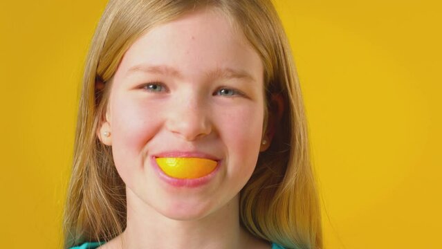 Studio Portrait Of Girl Using Orange Segment For Mouth And Teeth Against Yellow Background