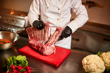 Professional chef cutting raw bbq beef ribs on the background of vegetables. Chief chef preparing...