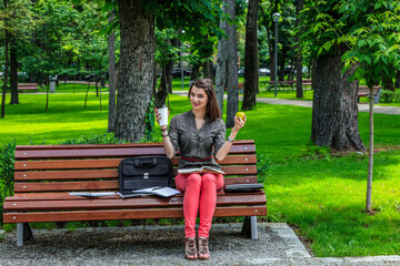 Fototapeta premium Young woman studying outside in a park holds a green apple and a cup of coffee looking to the camera. 