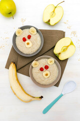 Funny cute kids childrens baby's healthy breakfast lunch oatmeal porridge in bowl look like bear face decorated with apple, banana, dried berry fruits. dessert food art on white wooden table