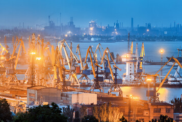 Sea commercial port and steel plant at night in Mariupol, Ukraine before the war. Industrial landscape. Cargo freight ship with working cranes bridge in sea port at dusk. Cargo port, logistic.