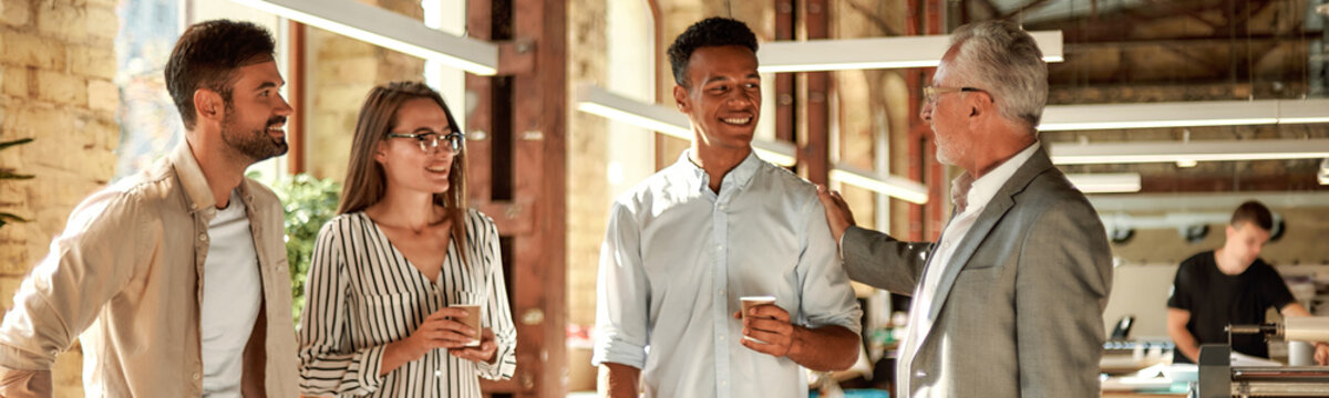 Good Job! Smiling Mature Man In Formal Wear Patting His Young Afro American Colleague On Shoulder While Standing With Coworkers In The Modern Office