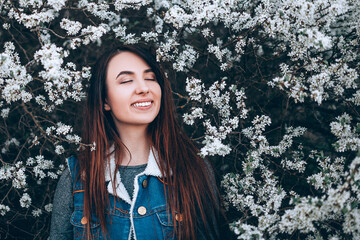 Beautiful girl stands with closed eyes and smiling on a background of flowering spring trees. The girl inhales the scent of spring flowers. Copy paste background