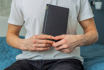 A man is sitting on the sofa and holding a book. A businessman dressed in a white T-shirt is sitting on the sofa and holding a black book. A black book in the hands of a man, a selective focus.