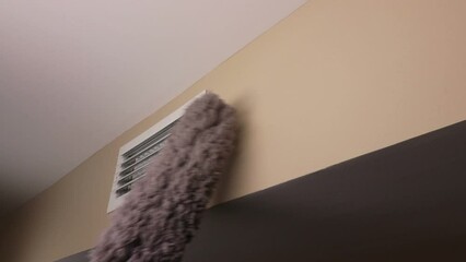Small white rectangle furnace vent grill being dusted by a male hand with a long gray dusting wand. Wall air duct vent near a ceiling being dusted with a long gray dust wand by an adult male hand.