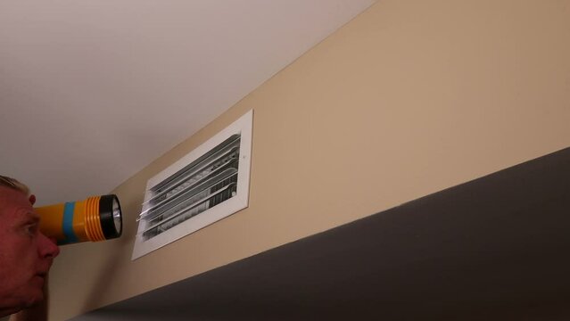 Mature male looking into a small home rectangle wall air duct vent with a flashlight to examine the quality. Home owner inspecting inside a small air duct vent with a flashlight for any maintenance.