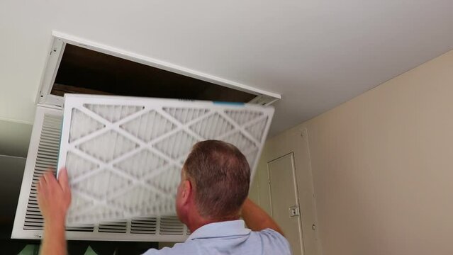 Adult caucasian male removes air filter and looks into the home furnace air intake vent for cleanliness. White guy takes out air filter from home furnace and examines the inside with a flashlight.