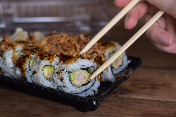 Persons hand taking crispy dragon roll sushi with chopsticks from take out container