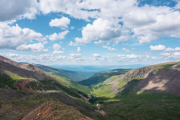 Naklejka premium Scenic aerial view from mountain pass to green forest valley among mountain ranges and hills on horizon at changeable weather. Green landscape with sunlit mountains under cumulus clouds in blue sky.
