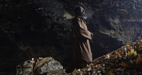 Hermit man with robe in the entrance of a old cave in front of a valley