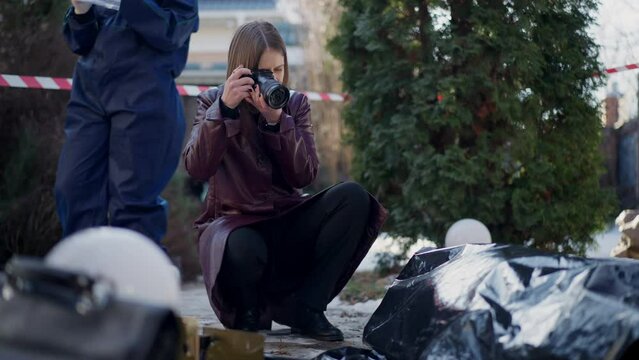Young focused police detective taking photos on crime scene with unrecognizable criminologist standing at background. Portrait of concentrated Caucasian woman photographing clues on winter day