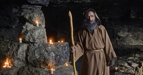 Medieval monk, with a staff in a hooded place, stands in a dark cave with glowing candles in the...