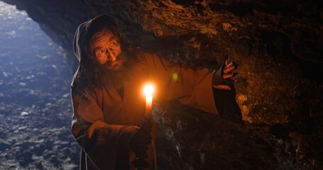 Old druid walks carefully in dark ancient cave with candle