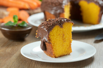 Close up piece of Brazilian carrot cake with chocolate frosting on wooden table with carrots in the background