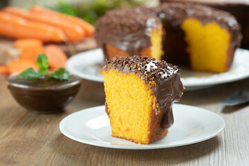 Close up piece of Brazilian carrot cake with chocolate frosting on wooden table with carrots in the background