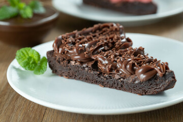 Piece of Chocolate Brownie Slice with Strawberry and Chocolate Icing. Wooden table with mint and chocolate chips in the background