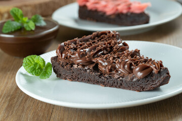 Piece of Chocolate Brownie Slice with Strawberry and Chocolate Icing. Wooden table with mint and chocolate chips in the background