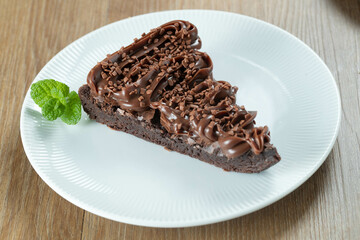 Piece of Chocolate Brownie Slice with Strawberry and Chocolate Icing. Wooden table with mint and chocolate chips in the background