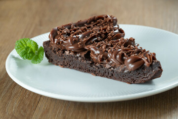 Piece of Chocolate Brownie Slice with Strawberry and Chocolate Icing. Wooden table with mint and chocolate chips in the background