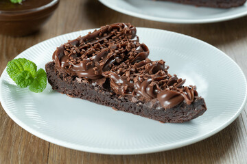 Piece of Chocolate Brownie Slice with Strawberry and Chocolate Icing. Wooden table with mint and chocolate chips in the background