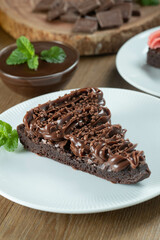 Piece of Chocolate Brownie Slice with Strawberry and Chocolate Icing. Wooden table with mint and chocolate chips in the background