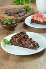 Piece of Chocolate Brownie Slice with Strawberry and Chocolate Icing. Wooden table with mint and chocolate chips in the background
