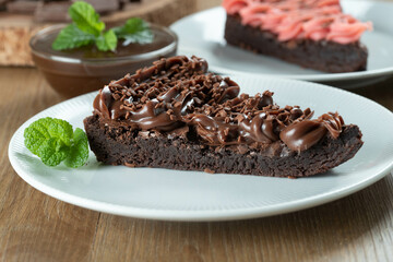 Piece of Chocolate Brownie Slice with Strawberry and Chocolate Icing. Wooden table with mint and chocolate chips in the background