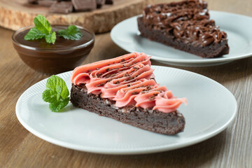 Piece of Chocolate Brownie Slice with Strawberry and Chocolate Icing. Wooden table with mint and chocolate chips in the background