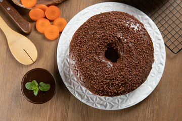 Brazilian carrot cake with chocolate frosting on wooden table with carrots in the background. Top view.